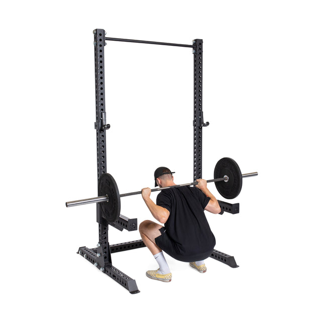 A person in black athletic wear and a cap performs a low bar squat at the bottom position inside a home gym, using the Bells of Steel Manticore Squat Stand Builder (3" x 3", 1" Holes) with a loaded barbell.