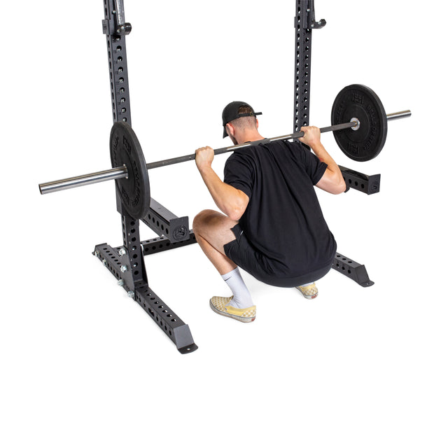 A man in black workout gear performs a deep squat with a barbell inside the Bells of Steel Manticore Squat Stand - Prebuilt (3" x 3", 1" Holes), viewed from behind against a white background—perfect for your home gym.
