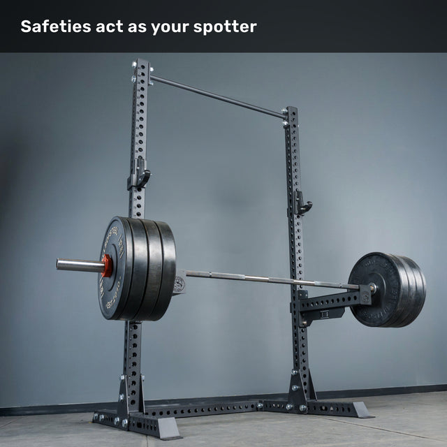 A loaded barbell rests on the safety pins of the sturdy Bells of Steel Manticore Squat Stand - Prebuilt (3" x 3", 1" Holes), with the text “Safeties act as your spotter” displayed at the top—a perfect setup for any serious home gym.