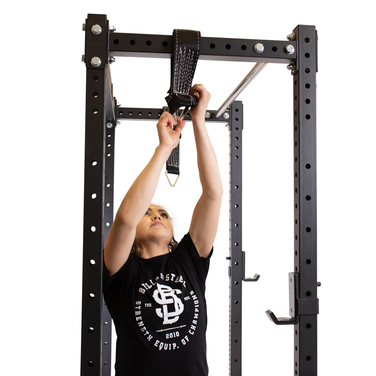 In a sleek home gym, a person in a black T-shirt expertly attaches Bells of Steel's Suspension Spotter Straps And Bodyweight System to the pull-up bar on a sturdy black metal power rack for invigorating suspension training.