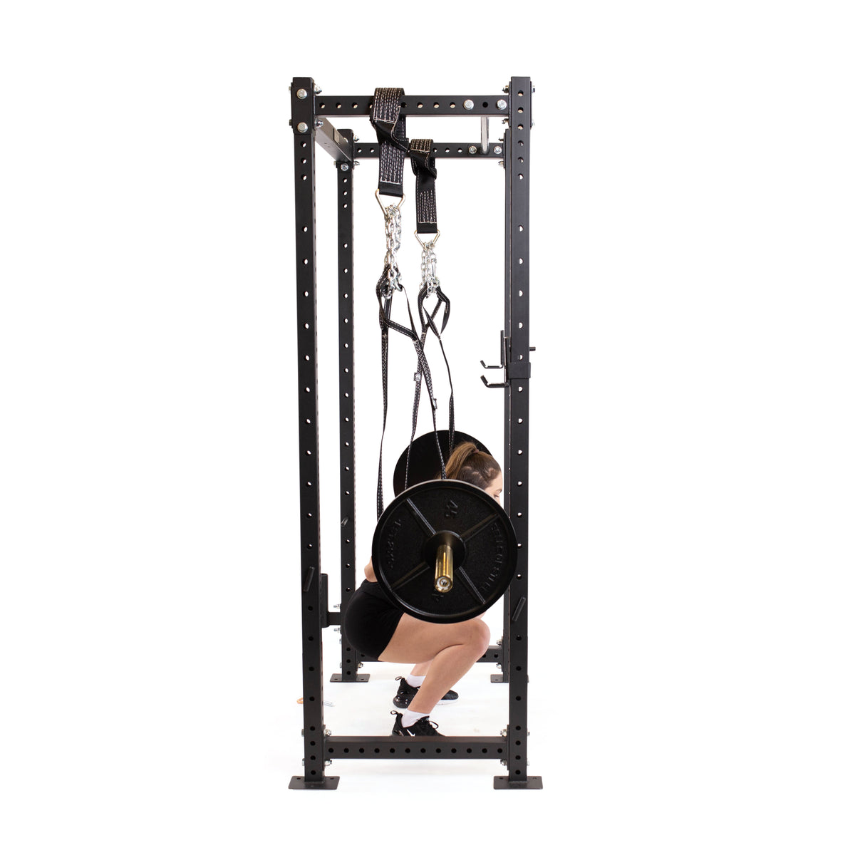 In a home gym, a person performs squats using Bells of Steel's Suspension Spotter Straps and Bodyweight System, with the barbell on their shoulders inside a power rack. Dressed in athletic wear, they appear focused and determined in their workout routine.