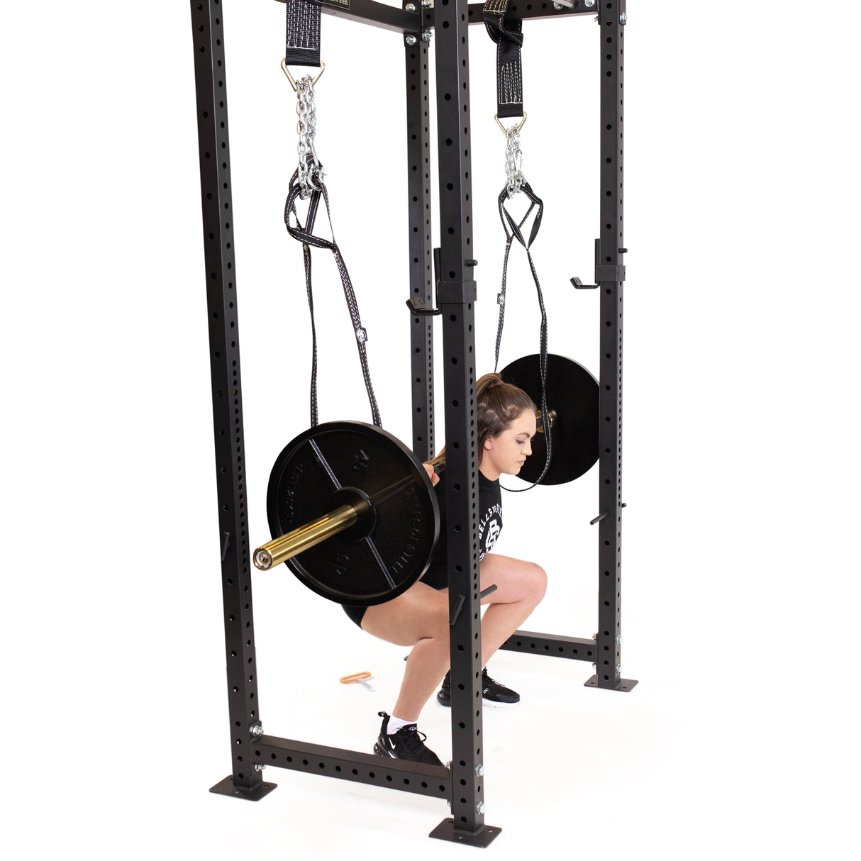In a home gym, a person with a prosthetic left leg performs squats using Bells of Steel's Suspension Spotter Straps and Bodyweight System in the power rack. Wearing black attire, they skillfully manage the barbell weights with safety spotter straps for support during their workout.
