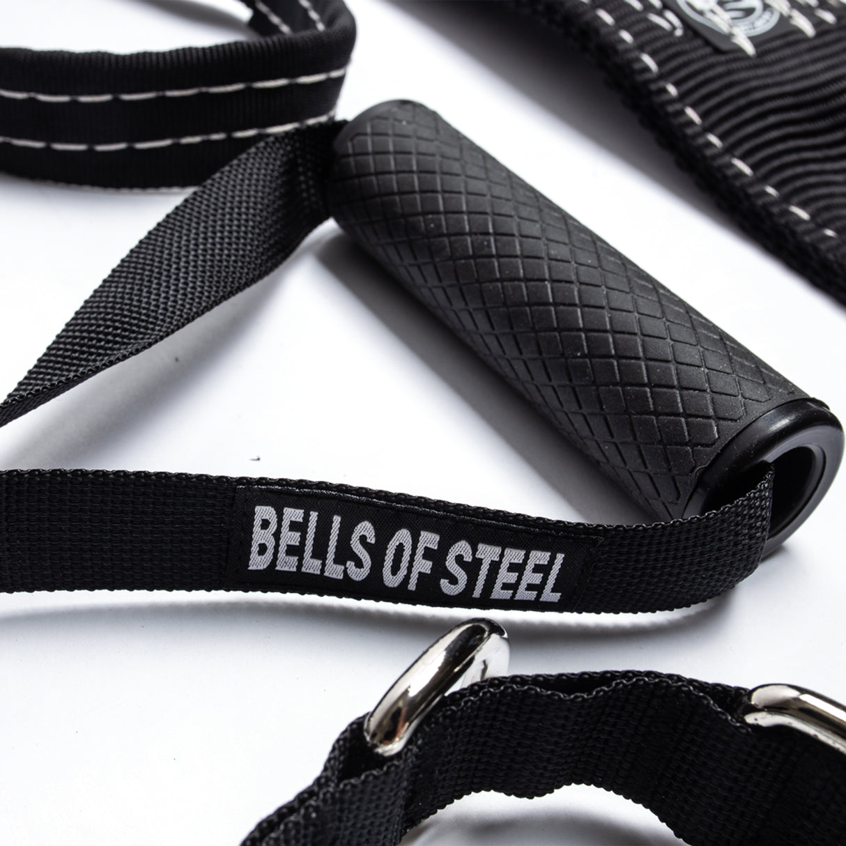 Close-up of a textured black exercise handle from Bells of Steel's Suspension Spotter Straps and Bodyweight System, perfect for home gym setups. The handle is attached to a strap with metal rings and stitching details, labeled "Bells of Steel," resting on a white surface.