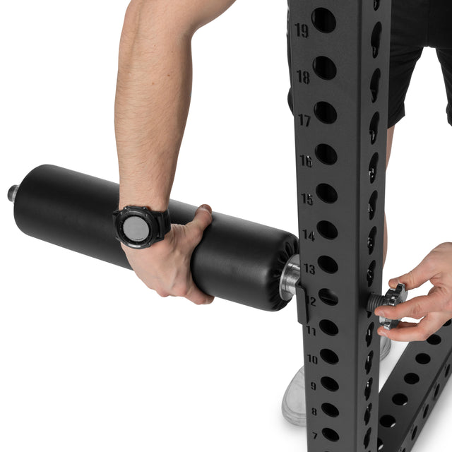 A person adjusts an exercise machine by inserting a Bells of Steel Split Squat Leg Roller Rack Attachment into the metal rack's numbered holes, securing it with a pin.
