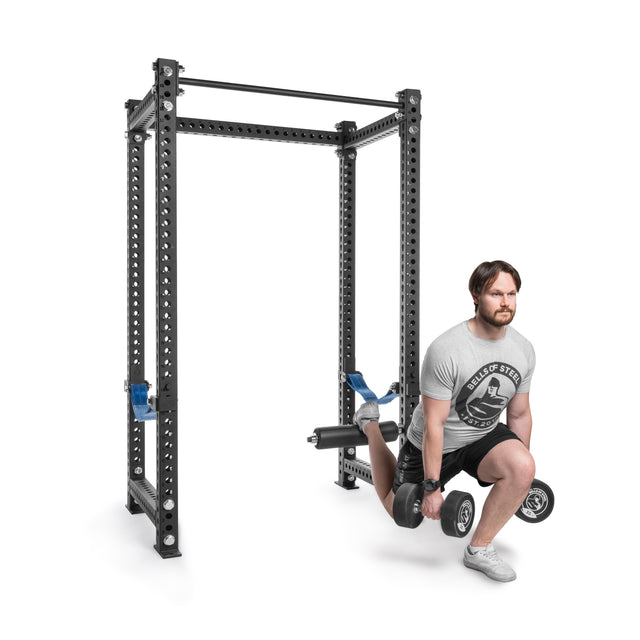A man in a light gray T-shirt, black shorts, and athletic shoes does a Bulgarian split squat using Bells of Steel's Split Squat Leg Roller Rack Attachment with adjustable blue safety arms in front of a large black squat rack.