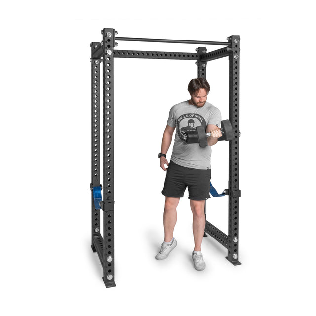 Wearing a gray T-shirt and black shorts, a man performs a bicep curl with a small dumbbell inside a power rack equipped with Bells of Steel's Split Squat Leg Roller Rack Attachment.