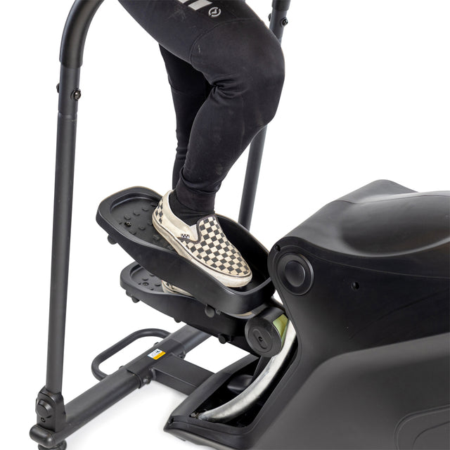 A person wearing black leggings and checkered slip-on shoes stands on a Bells of Steel Blitz Stepper, a low impact cardio stair climber. Only one leg and part of the machine are visible.