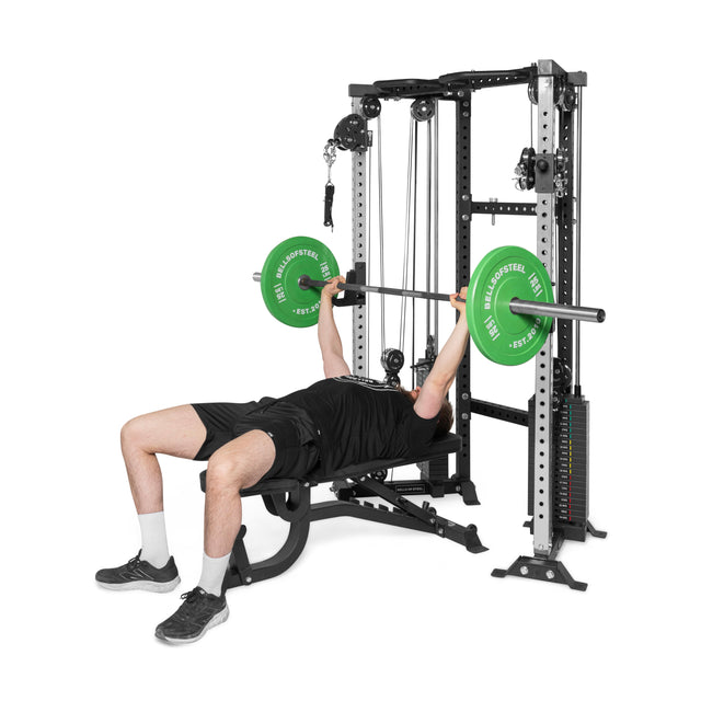 Someone wearing black rests on a gym bench, utilizing the Bells of Steel Functional Trainer Cable Tower to lift a barbell with green plates. The rubber mat flooring complements this versatile training equipment.
