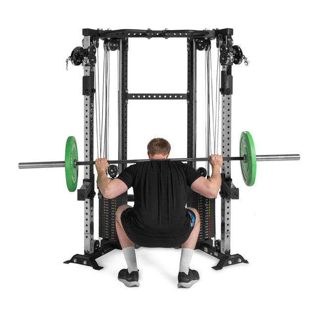 Dressed in black and facing away from the camera, a person squats with a barbell loaded with green plates inside a power rack, next to the Bells of Steel Functional Trainer Cable Tower.