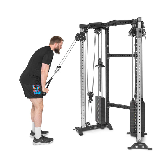 A bearded man in a black t-shirt and shorts confidently uses the Bells of Steel Functional Trainer Cable Tower at the gym, pulling down on its cables with both hands. The silver and black machine has neatly stacked weights at its center.