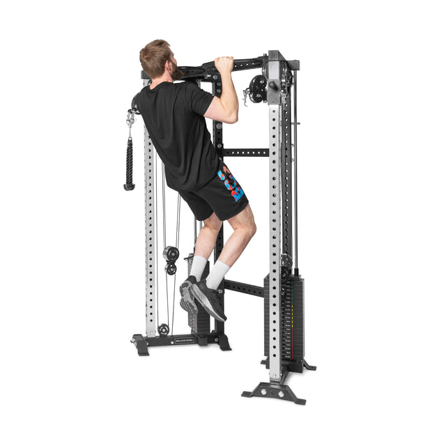 A person in a black shirt and shorts is doing pull-ups on a Bells of Steel Functional Trainer Cable Tower, using its adjustable weights and various attachments, including an overhead bar.