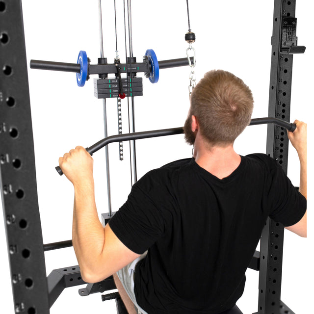 A bearded man clad in a black shirt intensely uses the Lat Pulldown & Low Row Rack Attachment by Bells of Steel, gripping the overhead bar on the Hydra & Manticore machine. He faces the apparatus with visible weights and pulleys against a stark white backdrop.