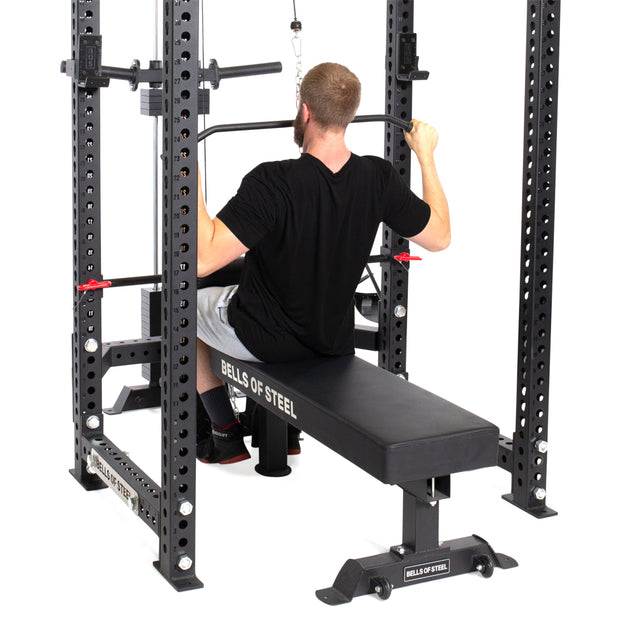 A man in a black shirt and gray shorts expertly uses the Bells of Steel Lat Pulldown & Low Row Rack Attachment - Hydra & Manticore, sitting on the bench with a firm grip on the wide bar above, showcasing his dedication amidst the bustling gym atmosphere.