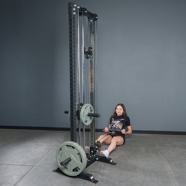 A woman prepares for a seated row in a gym, holding the Bells of Steel Fat Bar - Straight Handle Bar, which is attached to a cable machine with weight plates. The gym features gray walls and a tiled floor.
