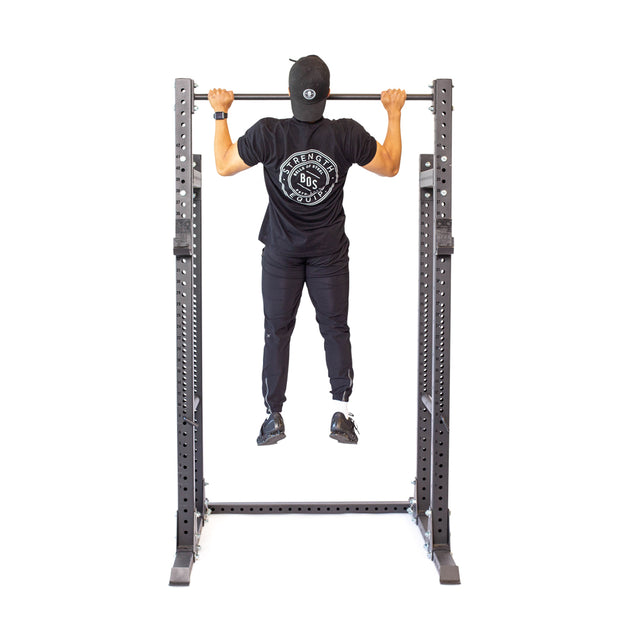 Wearing a black cap and clothes, an individual demonstrates grip strength on the Bells of Steel Straight Pull Up Bar, turning away from the camera against a white background.