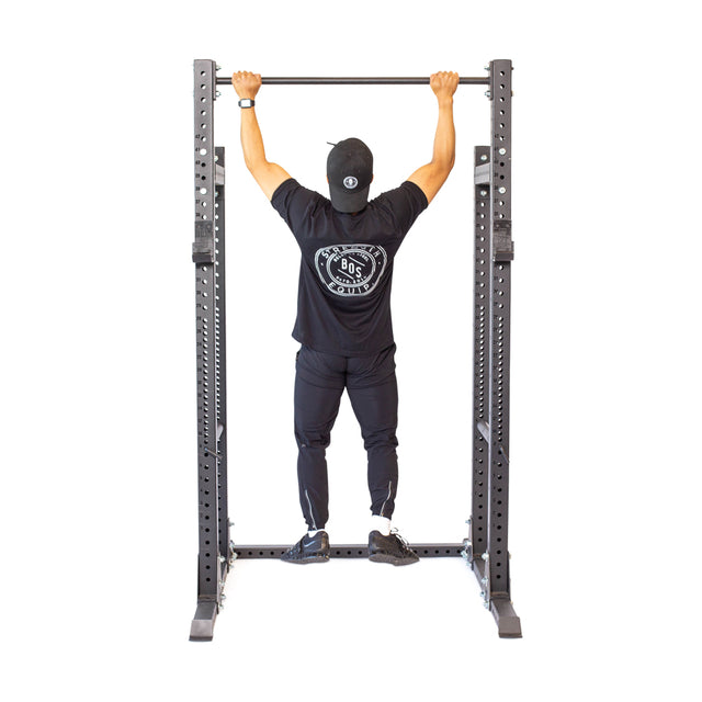 A person in a black outfit and cap performs a pull-up on the Bells of Steel Straight Pull Up Bar. The plain white background highlights the focus on the equipment and the individual's form.