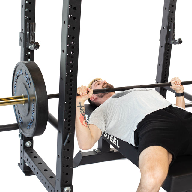 A man in a light gray shirt and black shorts prepares to bench press a barbell in a power rack, using Bells of Steel Sandwich J-Cups for secure support.