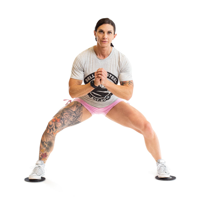 A woman in a gray t-shirt and pink shorts demonstrates a full-body lateral slide exercise with bent knees and clasped hands, using Bells of Steel Core Sliders against a white background.