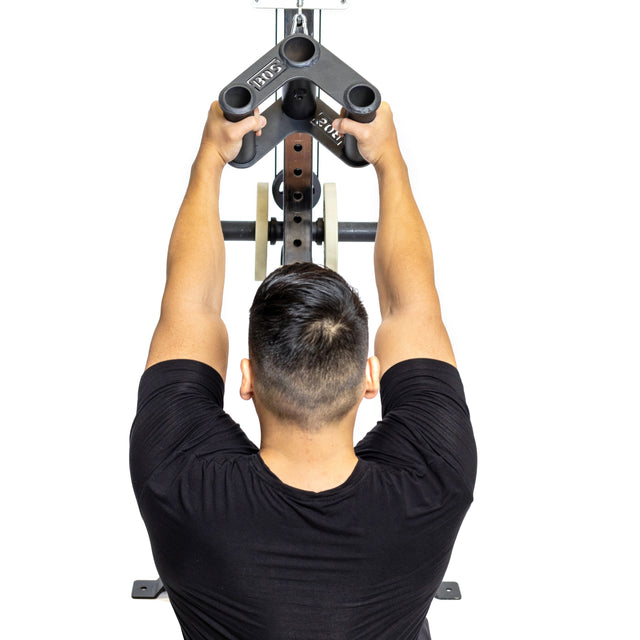 A person in a black shirt uses the Bells of Steel Fat Bar - T-Bar Row Cable Attachment at the gym, viewed from behind. Gripping the handles, they focus on enhancing upper body strength and grip during their intense workout session.
