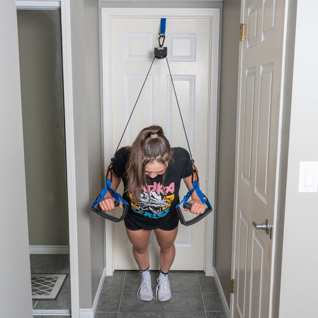 A person uses the Bells of Steel USA Rotational Pulley anchored above a doorway, leaning forward with their arms extended and feet together on a tiled floor, engaging in a challenging bodyweight workout.