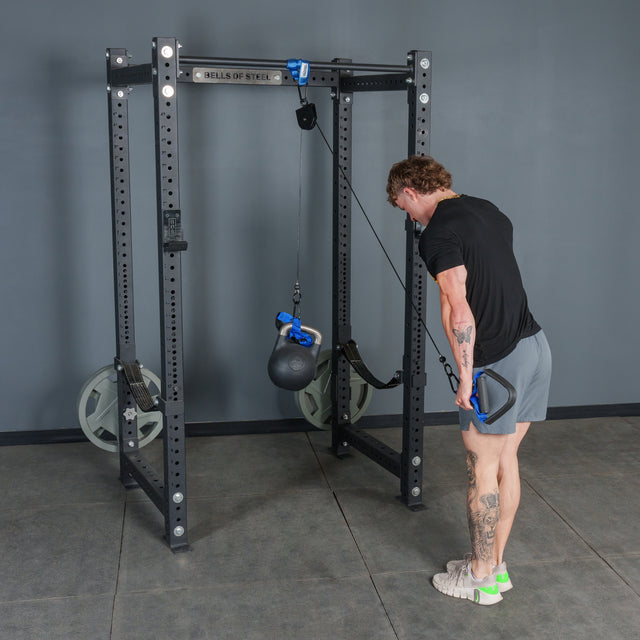 Wearing athletic attire, a person performs a triceps pushdown in a gym with gray floors and walls, using the Bells of Steel USA Rotational Pulley kit with a kettlebell attached.