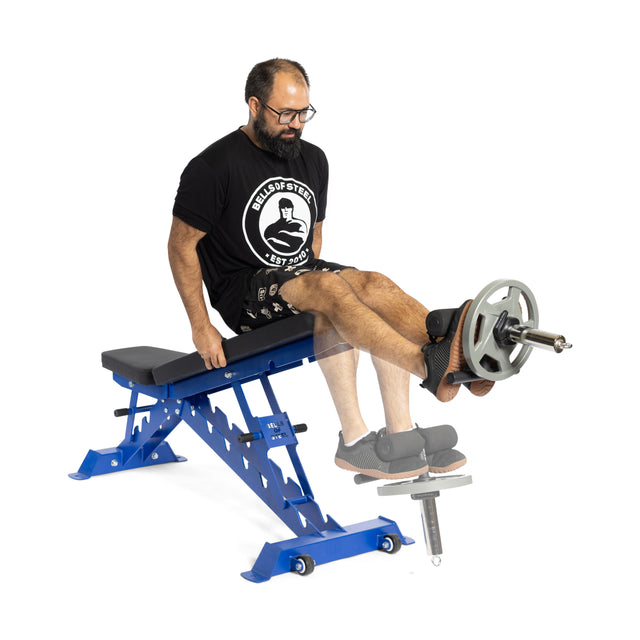 A bearded man in a black shirt uses the Bells of Steel Tibialis Trainer Bar on a blue adjustable bench, performing leg extensions with an Olympic weight plate for strength training and knee injury prevention.