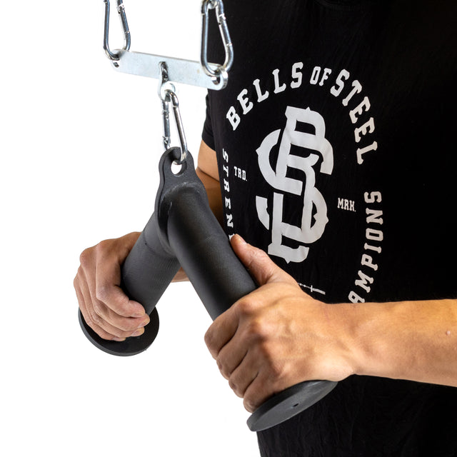 A person in a Bells of Steel shirt demonstrates grip strength using the Fat Bar - Tricep Pressdown, holding the black handle while performing tricep pushdowns on a metal cable machine against a plain white background.