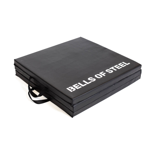 The Bells of Steel Trifold Mat is a black, square foam exercise mat with two side handles and "BELLS OF STEEL" in white on top. This durable crash pad conveniently folds, making it perfect for workouts and easy transport.