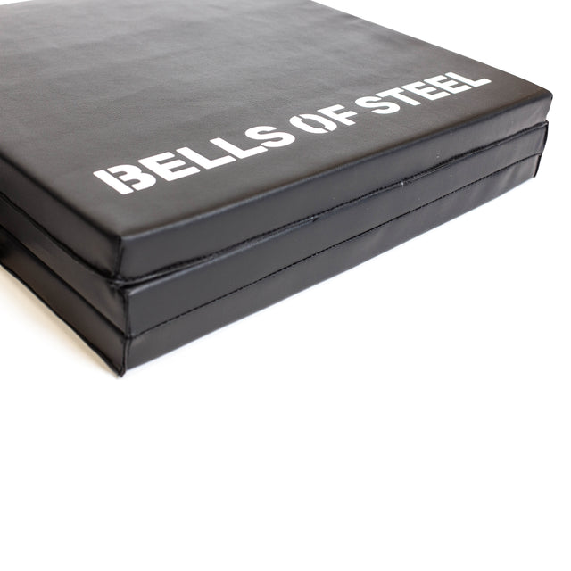 A close-up of the Bells of Steel Trifold Mat, a black, thick, non-slip workout mat with "BELLS OF STEEL" in white text. Designed for fitness or gym use, it offers comfort and support during exercises.