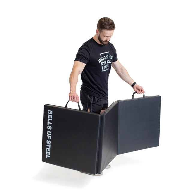 A man in a Bells of Steel shirt holds and unfolds a Trifold Mat by Bells of Steel. The foldable gym mat has handles, features non-slip material, and "BELLS OF STEEL" is printed on the side. The background is white.