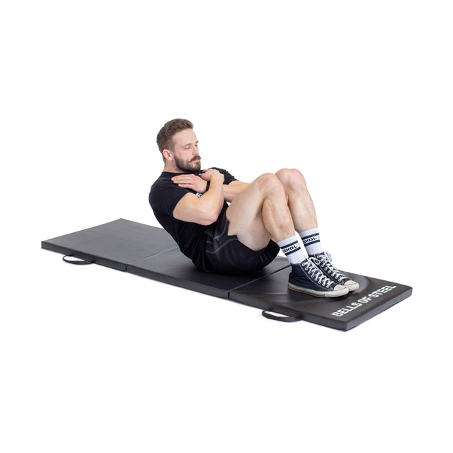 A man in athletic wear does crunches with crossed arms and bent knees on a black, non-slip Bells of Steel Trifold Mat.