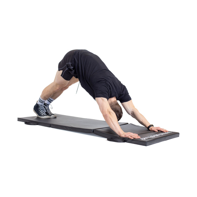 A person in black athletic wear and socks performs a downward dog pose on the Bells of Steel Trifold Mat against a white background.