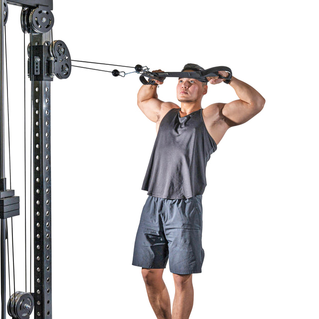A muscular man in a gray tank top and shorts uses the Bells of Steel RocIT Tricep Strap on a cable machine, performing a face pull by pulling the strap toward his face while standing against a white background.