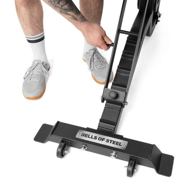 A person wearing gray sneakers and white socks adjusts the handle of a black Bells of Steel Adjustable Utility Bench, which has small wheels and multiple incline angles for customizable workouts.