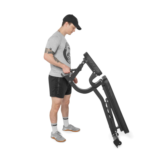 A man in athletic wear and a cap tilts the Bells of Steel Adjustable Utility Bench onto its wheels using the handle for easy transport. The plain white background highlights its portability and multiple incline options.