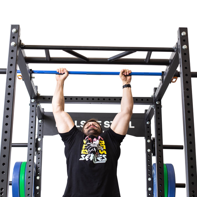 A man in a black graphic t-shirt does a pull-up on the Bells of Steel Above Rack Barbell Holder, attached to a power rack, viewed from below against a white background.