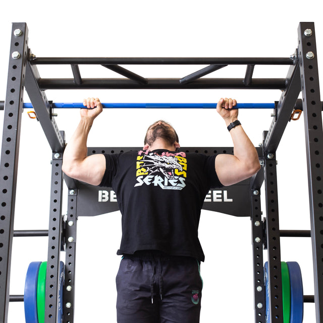 A man in a black graphic T-shirt and dark pants uses the Bells of Steel Above Rack Barbell Holder, performing a pull-up on a blue bar attached to a black power rack with weight plates, set against a white background.