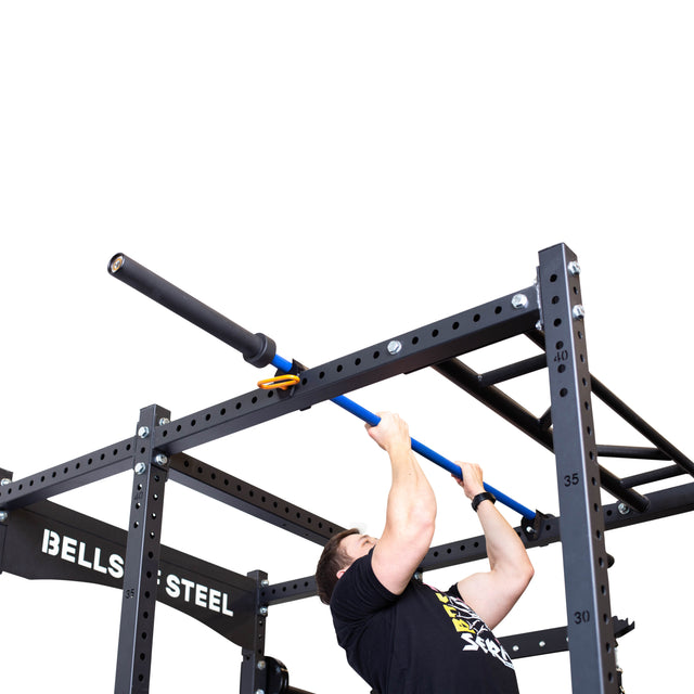 A person does a pull-up on a blue barbell held by the Above Rack Barbell Holder, secured to a black Bells of Steel power rack. The rack features the "BELLS OF STEEL" logo and stands against a white background.