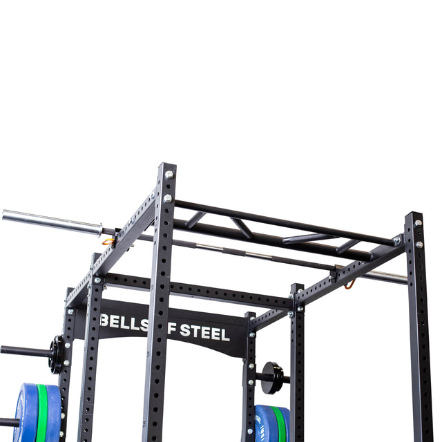 A close-up of the Bells of Steel Above Rack Barbell Holder attached to a black power rack with a pull-up bar and weight plates, displaying the "BELLS OF STEEL" logo on a crossbeam against a white background.