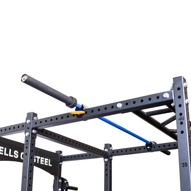 A black and blue barbell rests horizontally on a black power rack with numbered holes, held securely by yellow clips and the Bells of Steel Above Rack Barbell Holder. "Bells of Steel" branding is partially visible on the rack side.