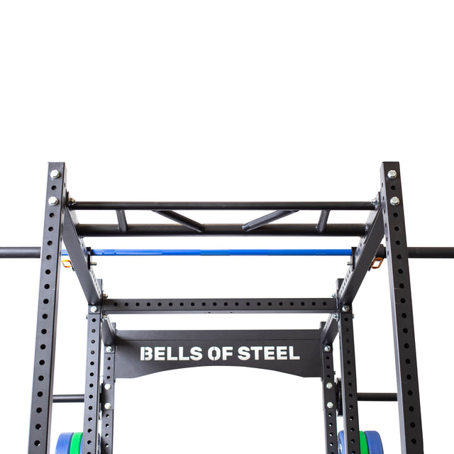 A close-up of a Bells of Steel black metal power rack with “BELLS OF STEEL” on the front, featuring pull-up bars, weight plate holders, and the Above Rack Barbell Holder attachment, set against a plain white background.