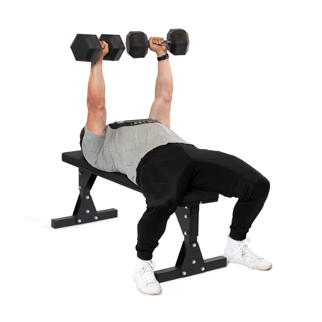 A person uses the Bells of Steel Flat Utility Bench for a dumbbell bench press, holding weights overhead. Dressed in a gray shirt, black pants, and white sneakers. Affordable and ideal for home gyms or budget-conscious users.