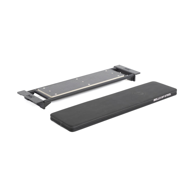 A rectangular black Bells of Steel Utility Seat for Hydra/Manticore and a separate steel textured mounting bracket, both shown on a white background. The pad features white "BELLS OF STEEL" text and fits any 3"x3" rack or adjustable bench.