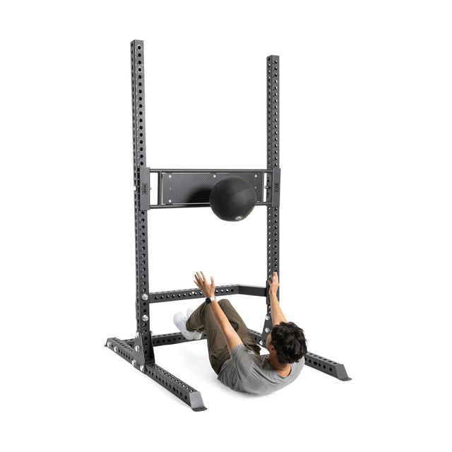 A user does sit-ups under a sturdy 3"x3" rack, tossing a black medicine ball at a horizontal target panel. Nearby is the Bells of Steel Utility Seat for Hydra/Manticore, adding workout versatility.