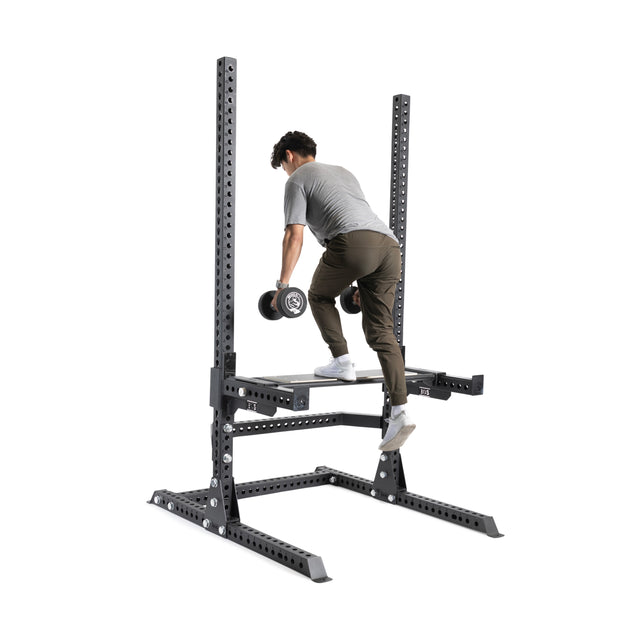 A person uses the Bells of Steel Utility Seat for Hydra/Manticore, stepping onto an elevated platform with a dumbbell in one hand, while wearing a gray shirt, brown pants, and white sneakers.