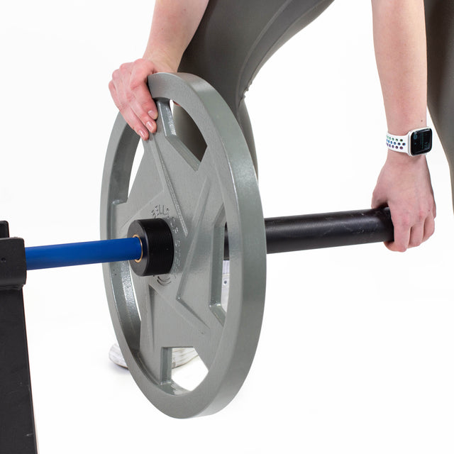 In a gym, someone in a gray outfit uses Gray Mighty Grip Olympic Weight Plates by Bells of Steel to attach to a black and blue barbell while wearing an Apple Watch. The ergonomic design ensures ease of use, and the minimalist white background highlights the focused intensity.