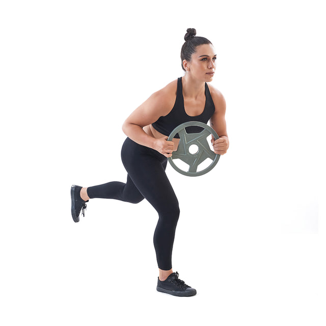 A woman in black athletic wear performs a fitness exercise, balancing on one leg while holding a Bells of Steel Gray Mighty Grip Olympic Weight Plate. Her hair is styled in a top bun as she stands against a plain white background.