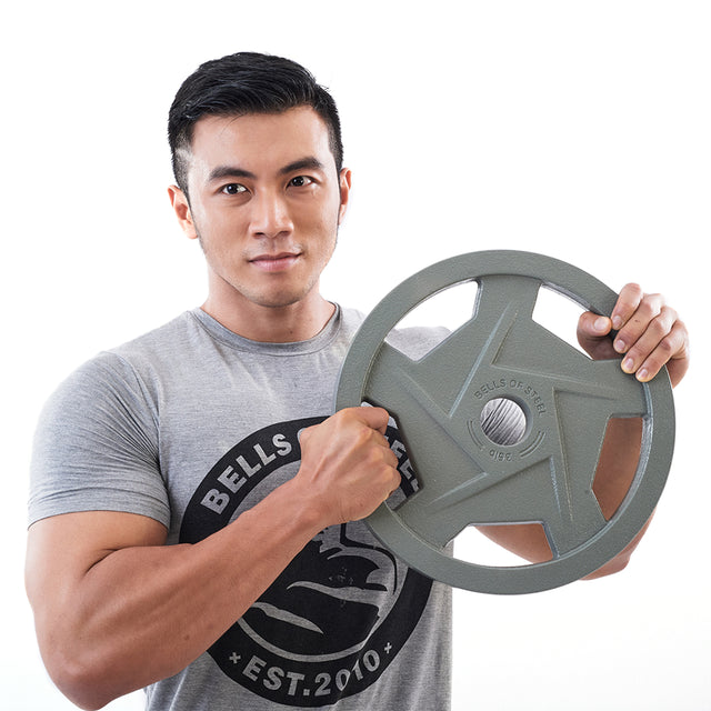 A person in a gray "Bells of Steel" t-shirt confidently poses against a white background, showcasing the ergonomic Gray Mighty Grip Olympic Weight Plate with a hammertone powder coat.