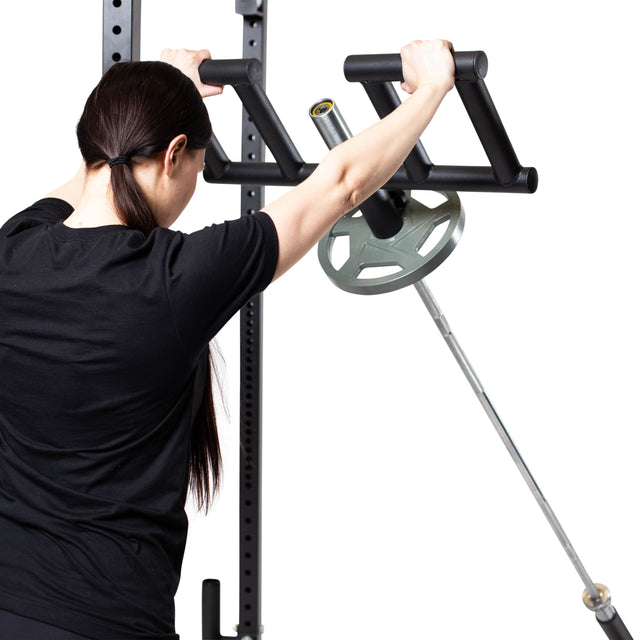A woman with long dark hair in a ponytail uses the Bells of Steel Viking Press Attachment with a loaded Olympic bar for a standing upper body exercise in the gym.