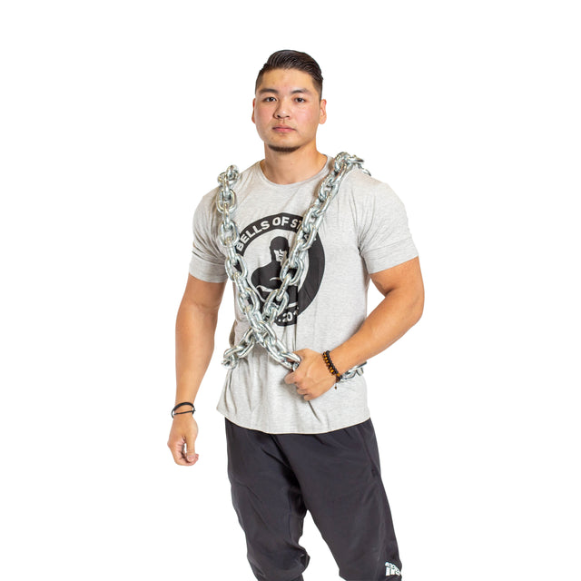 A man showcases the Bells of Steel Chain Weight Vest, standing against a plain white background in a gray t-shirt and black shorts, demonstrating the intensity of strength training with weighted chains draped across his body.